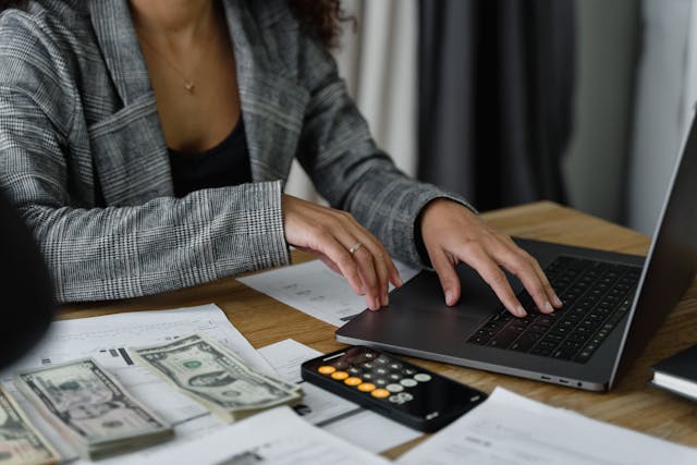 a woman in a suit typing on a computer while a calculator and stacks of cash are laid out in front of her